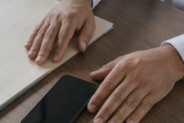 A clinician's hands resting on a closed paper chart, a phone lying flat beside them with a dark screen.